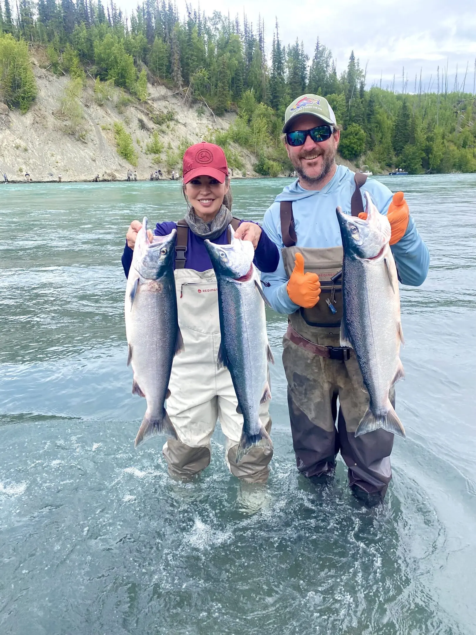 Two people holding large fish by river.