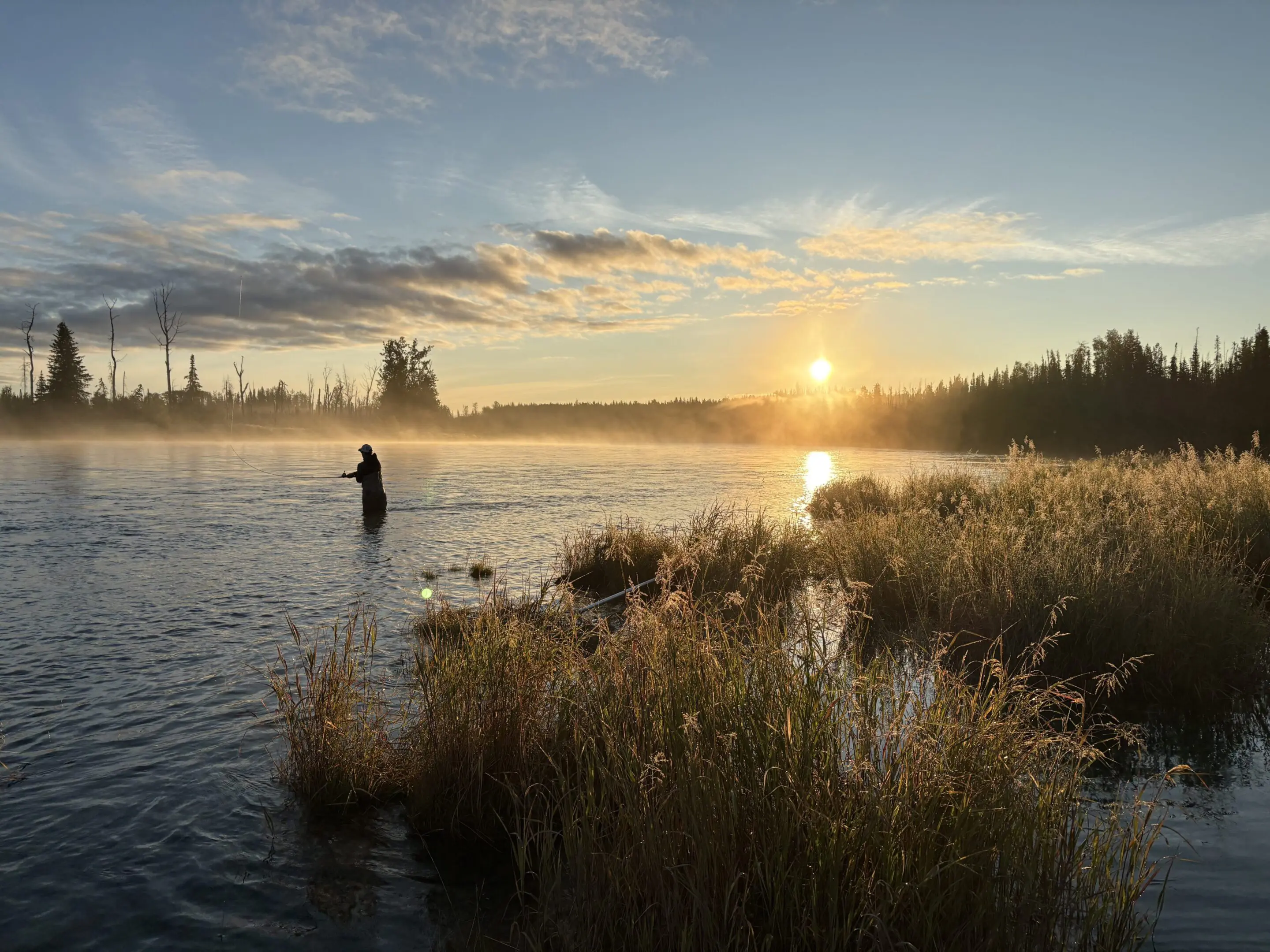 Person fishing at sunrise on misty river.